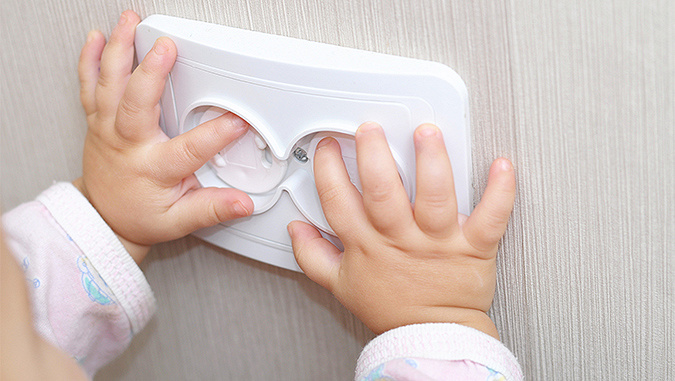 Les mains d'un enfant s'appuyent sur une prise électrique. Il essaye de mettre ses doigts à l'intérieur, mais la prise a des protections qui l'empêchent de le faire.