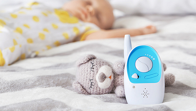 Sur un lit, un babyphone blanc-bleu de la marque Kinderkraft posé contre une peluche - ours en laine. Dans le fond, un bébé qui dort.
