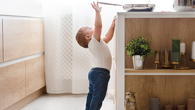 Un petit enfant habillé en jean et en chemisier blanc est debout dans la cuisine. Il grimpe sur la pointe des pieds et atteint avec sa poignée la casserole qui est sur la table. 
