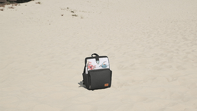 Pendant une journée ensoleillée, sur une plage de sable, un sac à dos blanc-gris de la marque Kinderkraft avec motif de fleurs et d'oiseaux.
