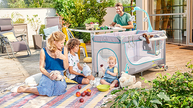 Sur une terrasse, une maman qui joue avec deux enfants. A côté, un parc pour bébé de la marque Kinderkraft avec un carrousel pour accrocher des jouets dessus, et un papa souriant dans le fond.