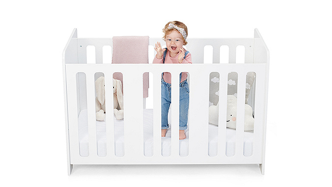 Un lit à barreaux de la marque Kinderkraft sur un fond blanc. Dans le lit une petite fille souriante et des peluches autour d'elle.