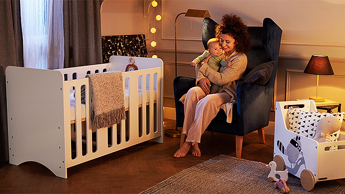 Le soir, dans une chambre d'enfant, une maman assise dans un fauteuil tient un petit enfant. A côté se trouve un parc pour bébé et un coffre à jouets Racoon de la marque Kinderkraft.
