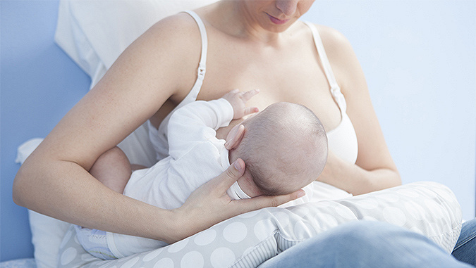 Une maman assise dans un lit, le dos contre le mur, nourrit le bébé allongé sur un oreiller spécial d'allaitement Kinderkraft. 