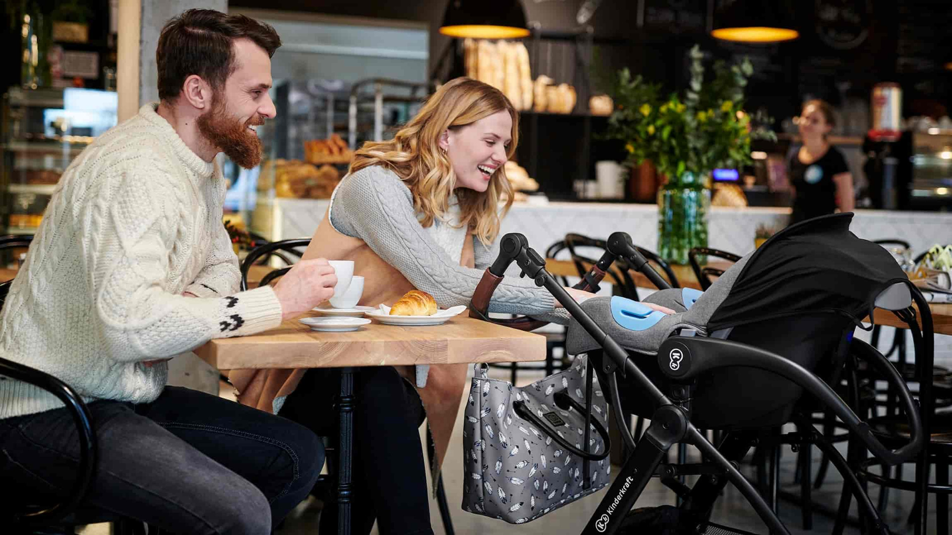 Couple rieur assis dans un café. La femme tend la main dans le chariot XMOOV. L'homme tient une tasse de café dans sa main.