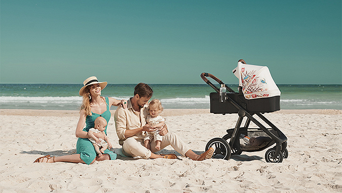 Lors d'une journée ensolleillée, des parents sont assis sur une plage au bord de la mer avec deux petits enfants. A côté, une poussette - nacelle de la marque Kinderkraft avec nacelle colorée. 