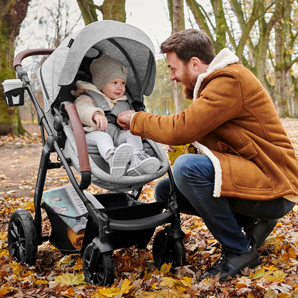 Un père en veste se penche vers un enfant assis dans une poussette. L'enfant rit, il y a des feuilles d'automne tout autour.